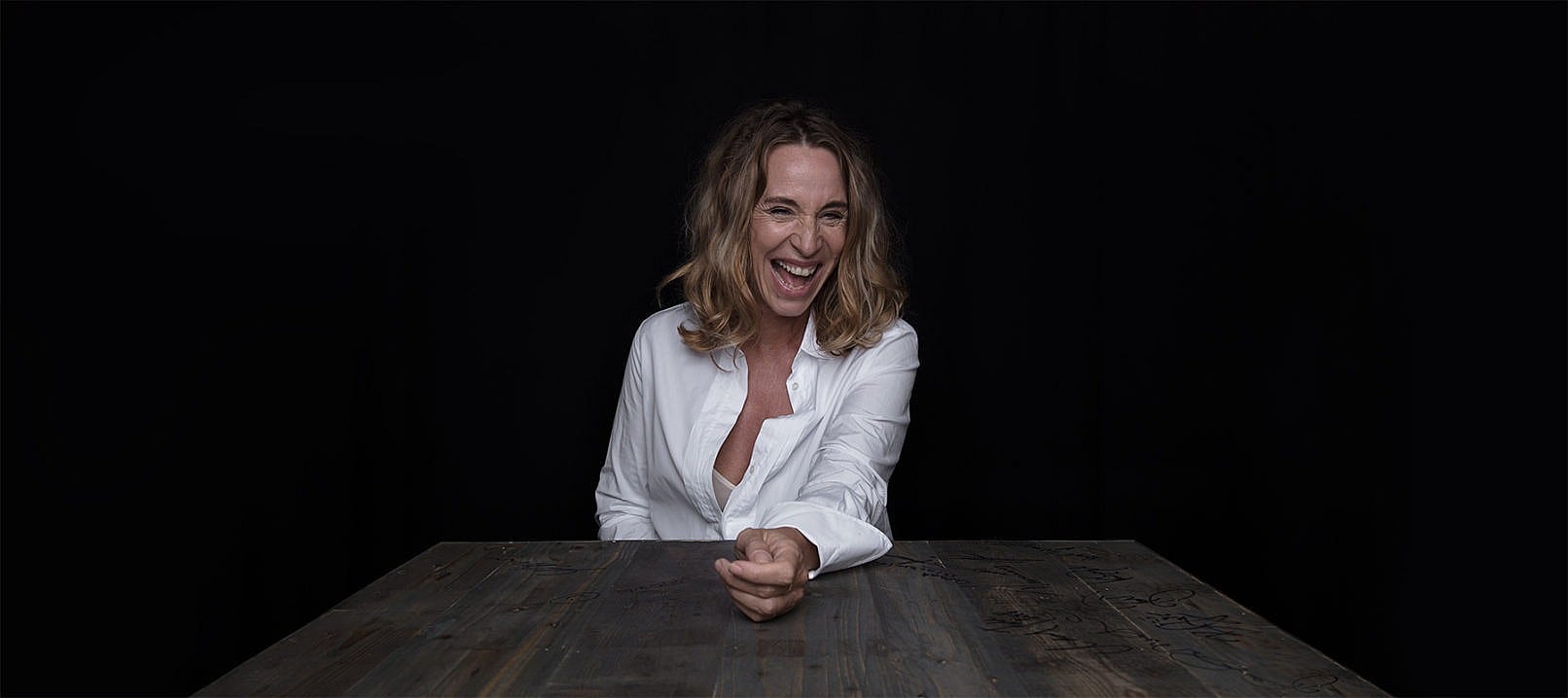 Ann Eleonora Jørgensen, sidder og griner ved et bord - Fotograf Christian Krog - Portrætfotograf
